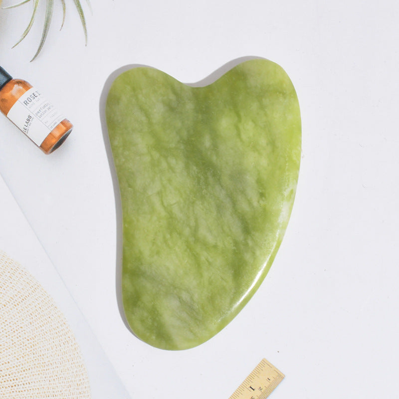 Heart-shaped jade gua sha stone for the face, shown flat beside a facial oil bottle and a ruler for scale