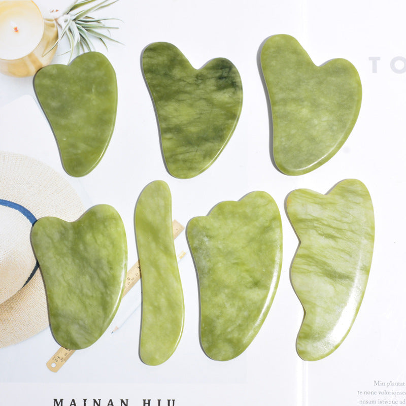Six jade gua sha stone shapes laid flat, including heart, curved board and notched jawline contours, cut from green phoenix jade
